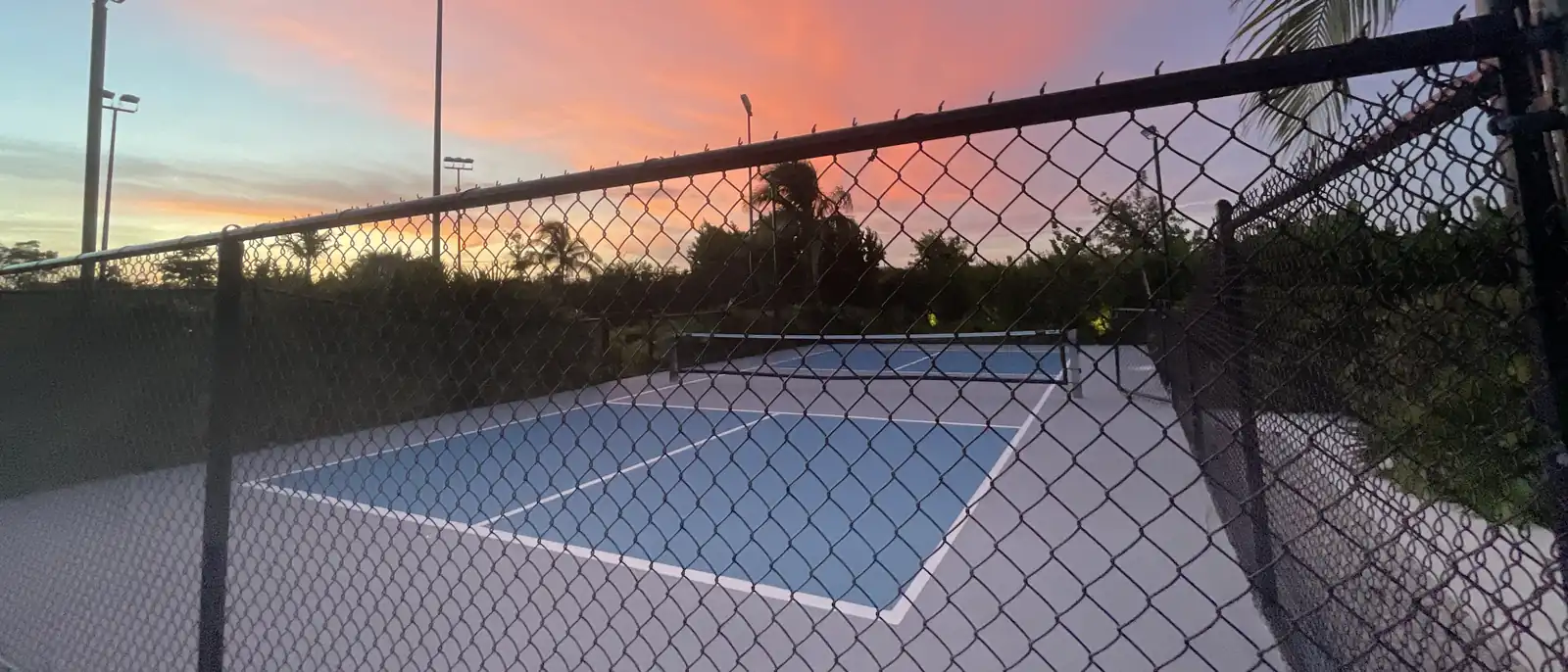 Finished pickleball court with net and regulation lines seen through the perimeter fence under the last light of the day